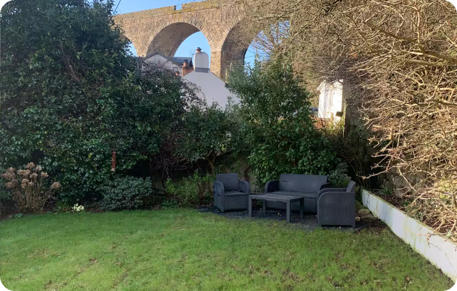 Garden seating area with black furniture near lush greenery and a historic stone viaduct in the background.