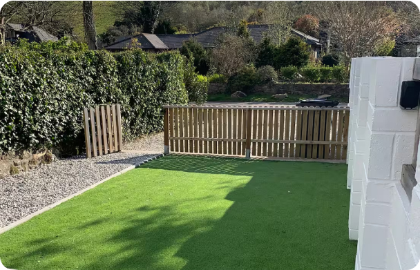 Small backyard with artificial turf, wooden fence, and gravel pathway; surrounded by lush greenery and trees.