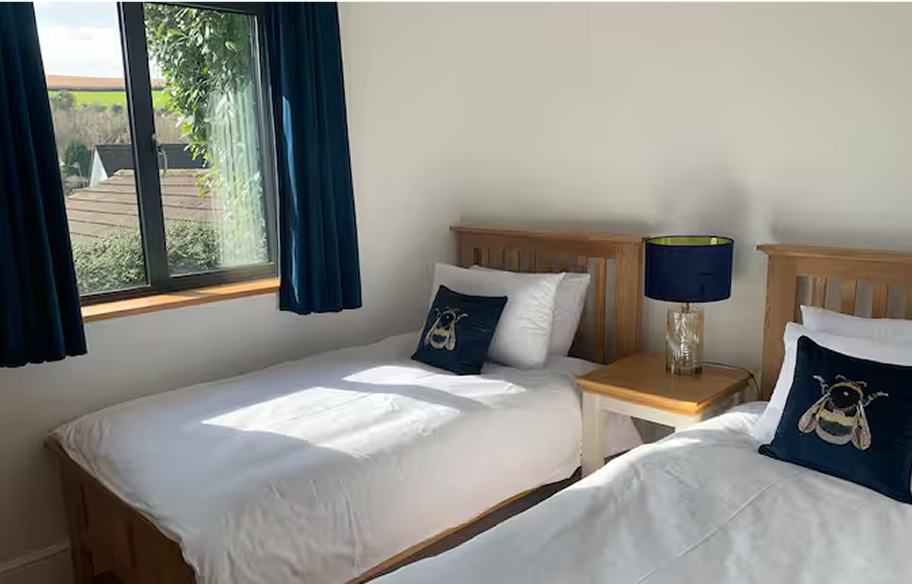 Bright twin bedroom with white bedding, wooden furniture, bee pillows, and view from window with blue curtains.