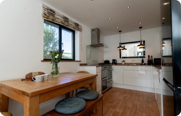 Modern kitchen with wooden dining table, cozy cushions, and sleek appliances under pendant lights.