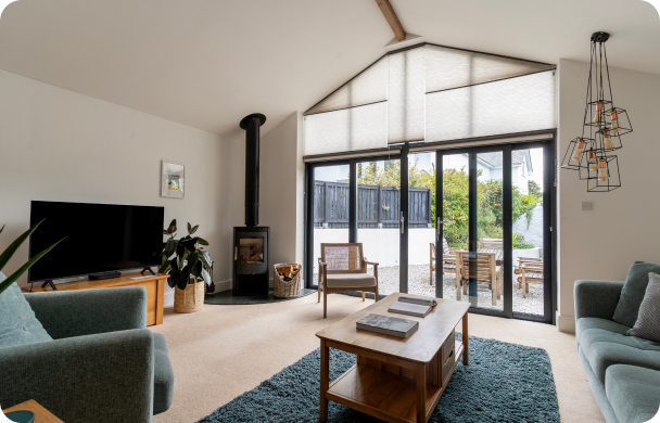 Modern living room with large windows, cozy sofas, a TV, and a fireplace, opening to a bright patio area.