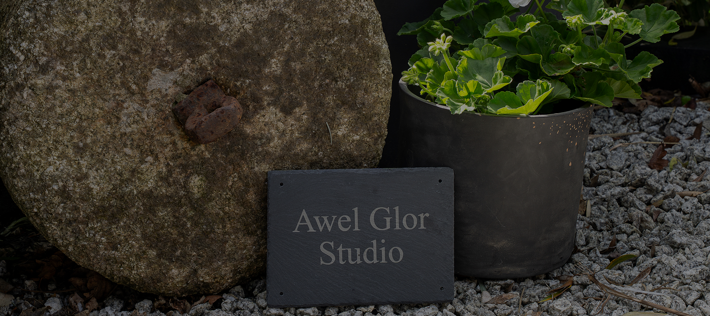 Stone mill with Awel Glor Studio sign and plant in pot on gravel path for rustic garden decor.