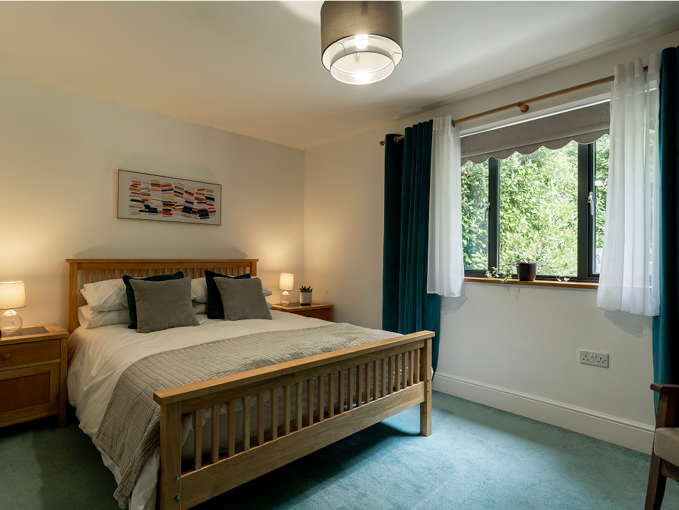 Cozy bedroom with wooden bed, green curtains, and large window view. Modern decor and soft lighting create a relaxing