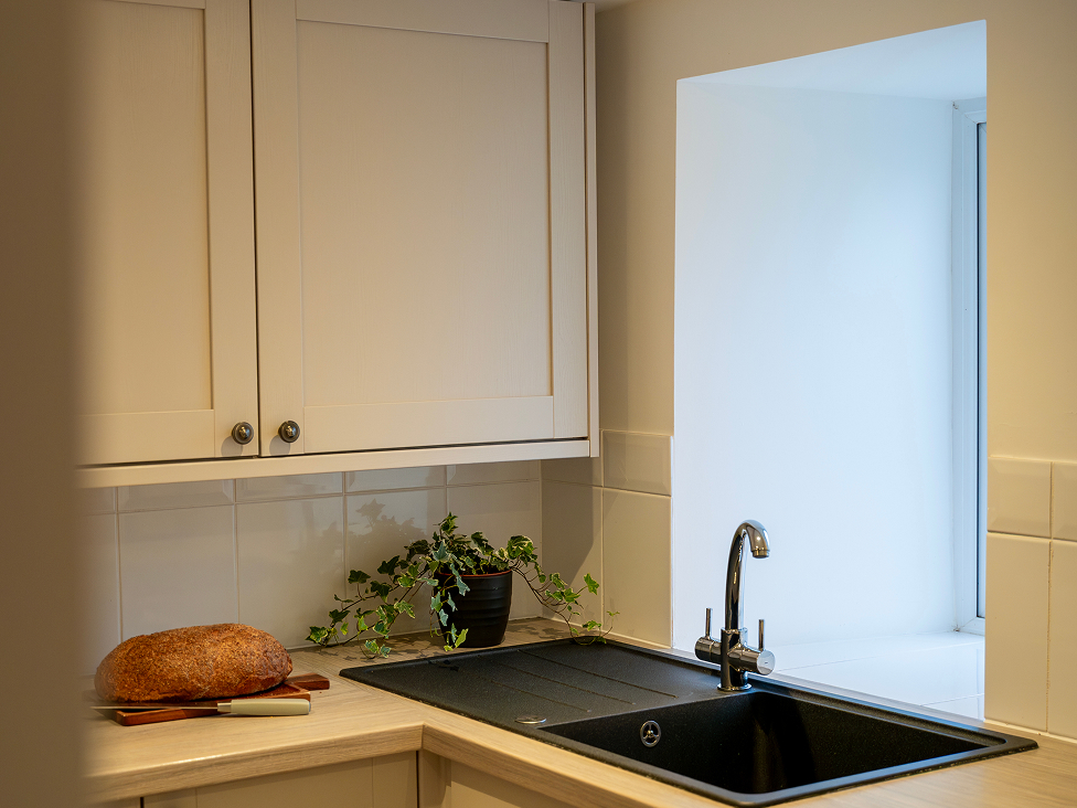Modern kitchen corner with beige cabinets, sleek black sink, fresh bread, and a green plant near a bright window.