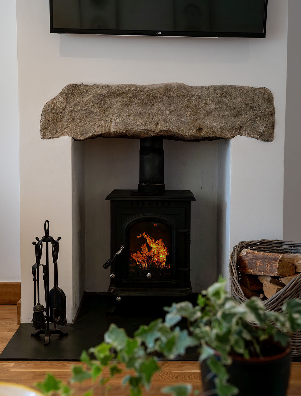 Cozy stone fireplace with a lit wood-burning stove, flanked by logs and tools, enhancing warm interior ambiance.