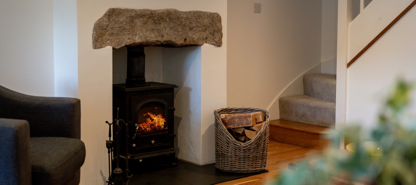 Cozy living room with wood-burning stove, stone mantel, and wicker basket of firewood beside carpeted stairs.