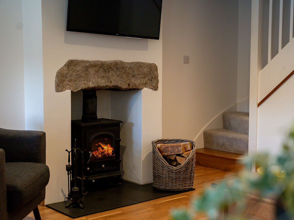 Cozy living room with wood-burning stove, basket of logs, mounted TV, and carpeted stairs.