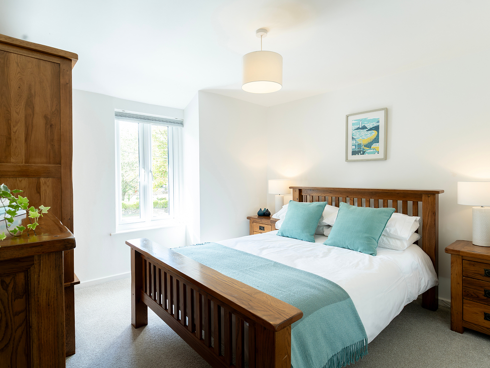 Cozy bedroom with wooden furniture, teal accents, large window, and white walls. Bright and inviting ambiance.
