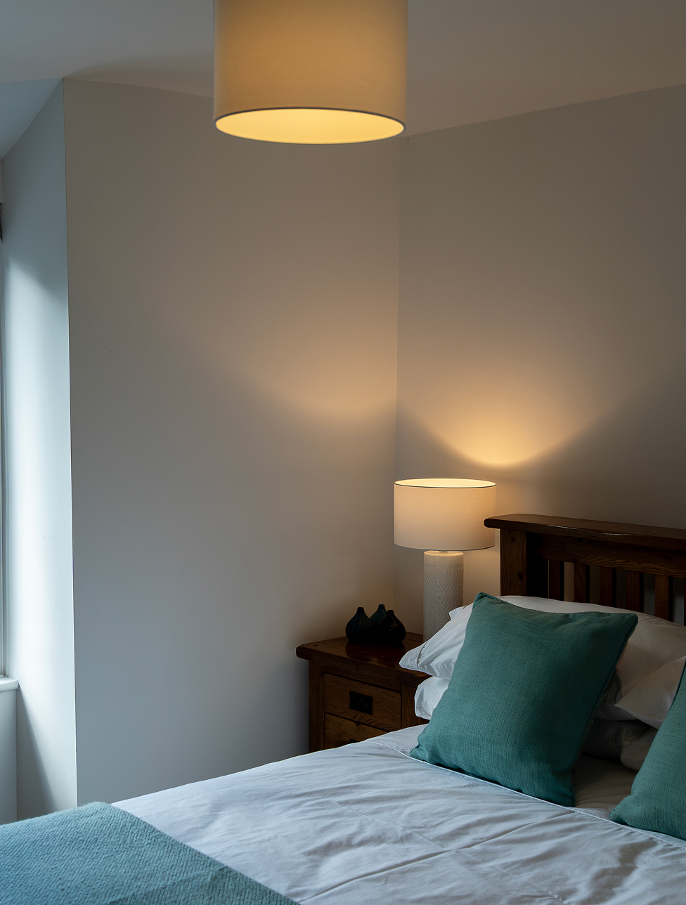 Cozy bedroom with teal cushions, wooden bed, bedside lamp, and natural lighting from a window.