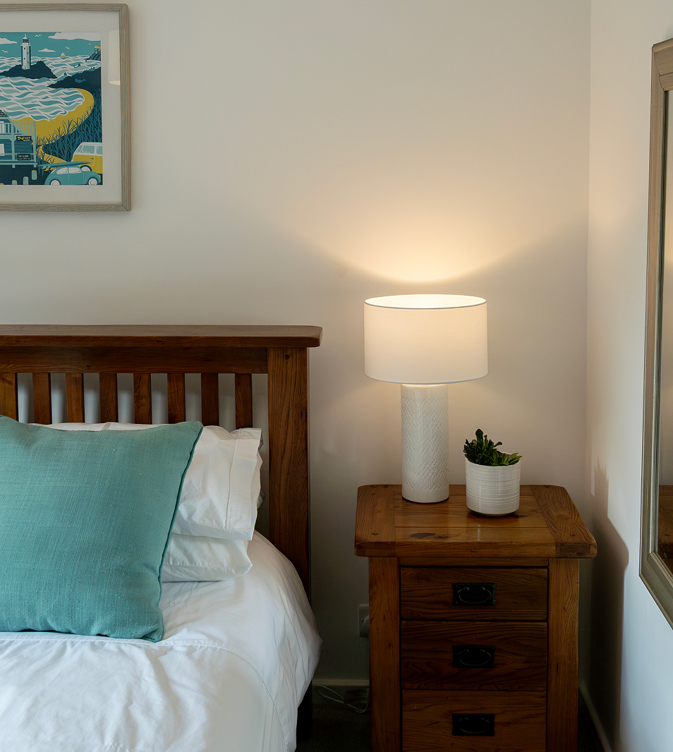 Cozy bedroom corner with wooden bed, teal pillow, nightstand, warm lamp light, and framed wall art.