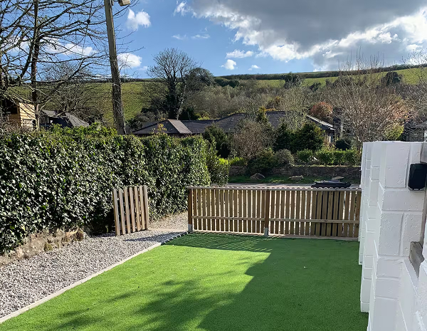 Sunny backyard with lush artificial grass, wooden fencing, and scenic countryside view under a partly cloudy sky.