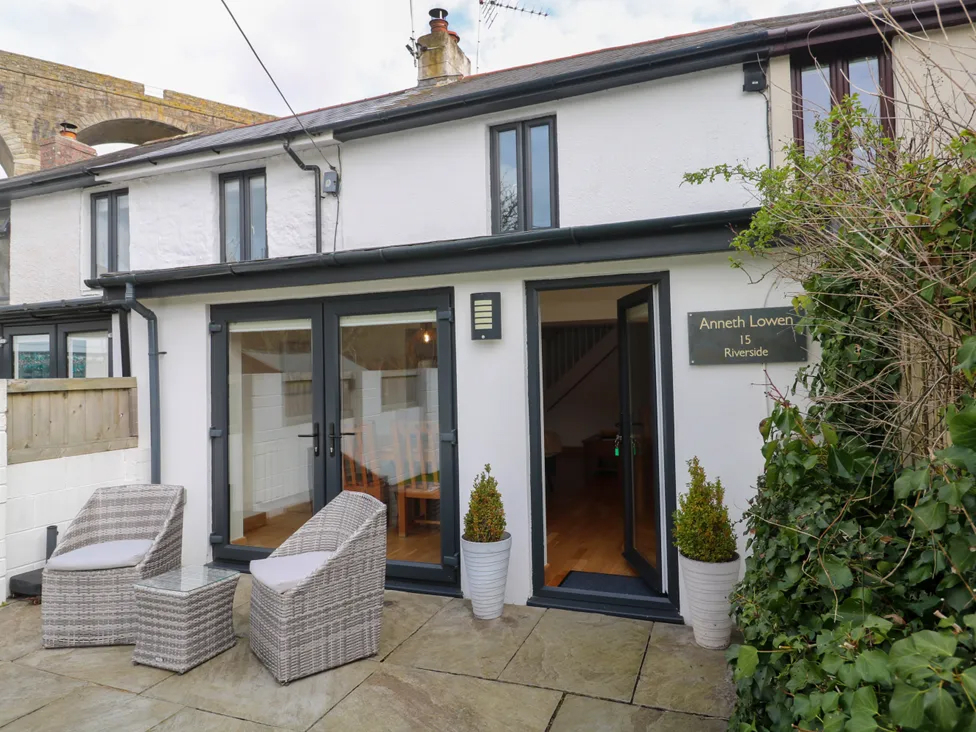 Cozy cottage patio with wicker chairs and glass doors, under a stone viaduct. Lush greenery surrounds the entrance.