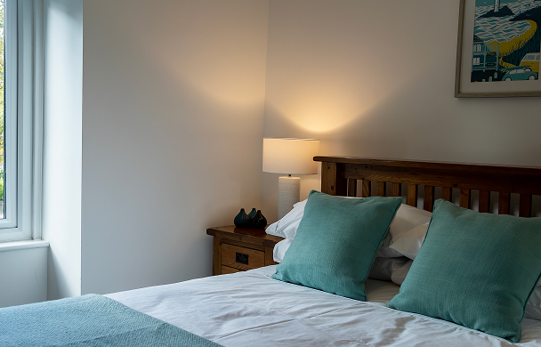 Cozy bedroom with teal pillows, wooden bed, bedside lamp, and natural light from a window.