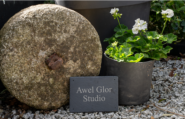 Stone wheel and potted flowers with Awel Glor Studio sign, set on gravel path.