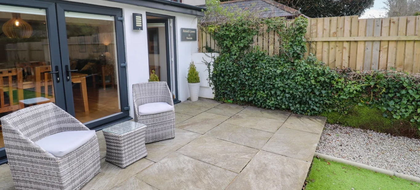 Modern patio with two wicker chairs and a table, surrounded by a lush green garden backdrop.