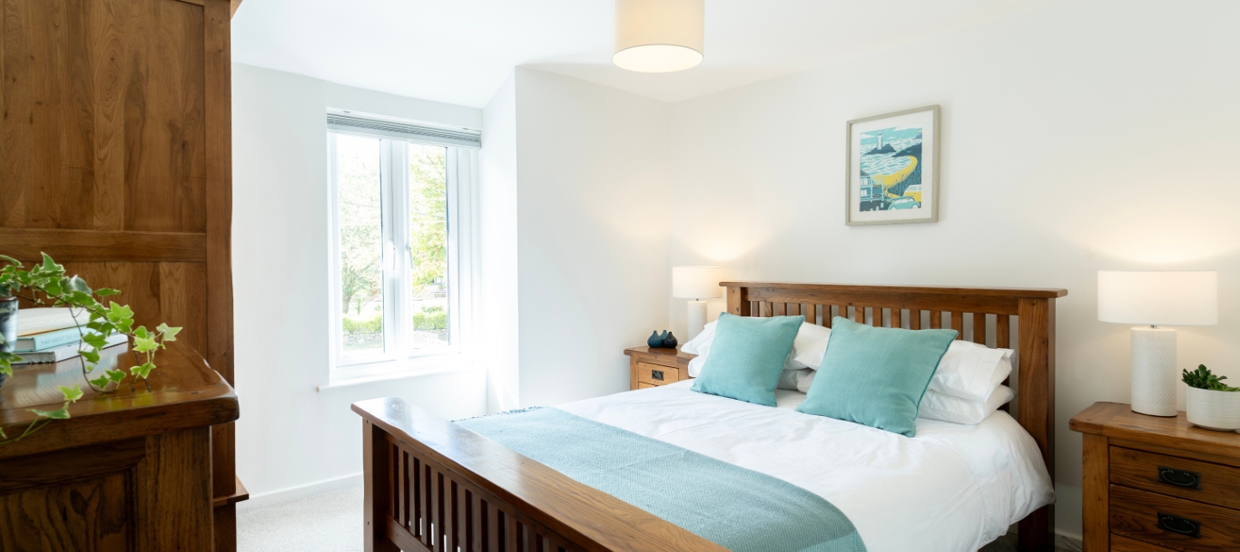 Cozy bedroom with wooden furniture, teal accents, large window, and a framed artwork above the bed.