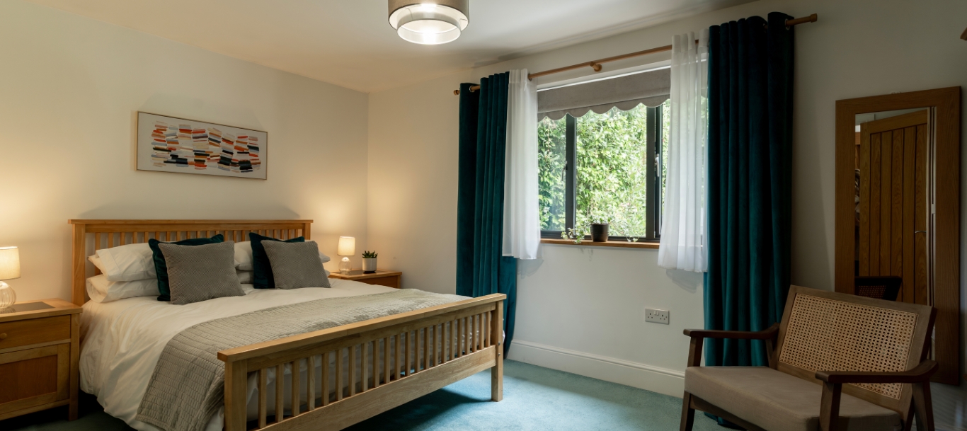 Cozy bedroom with wooden furniture, double bed, and large window with teal curtains, providing natural light and relaxation.