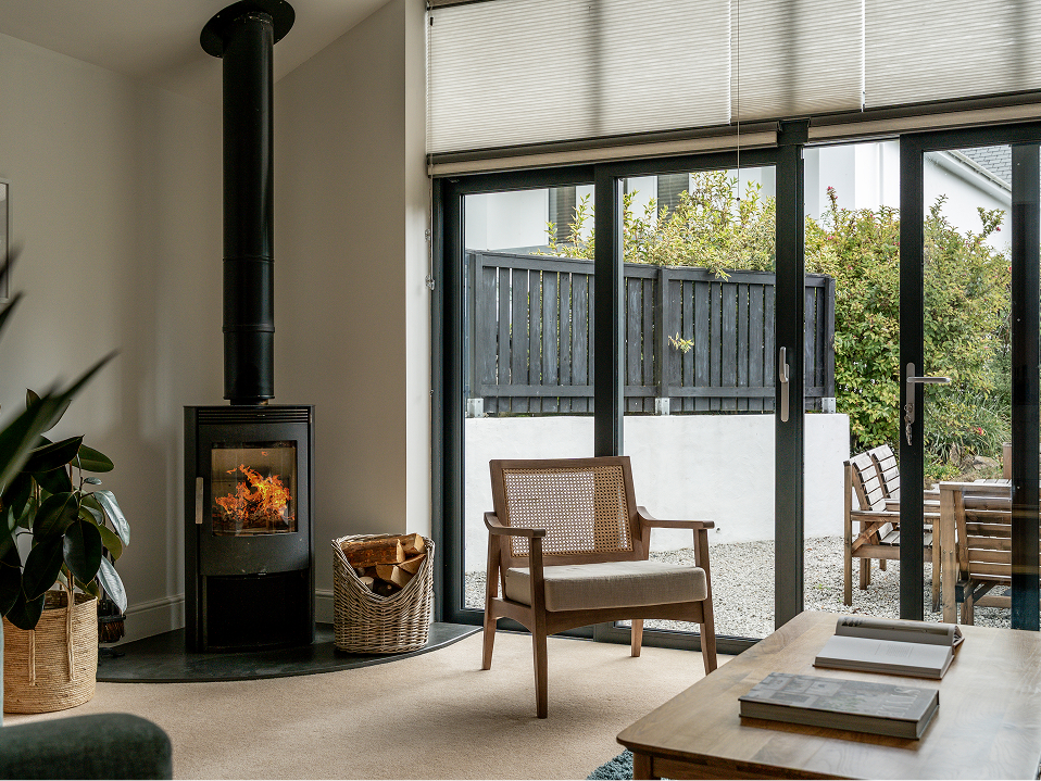 Cozy living room with a modern wood stove and wicker chair, featuring large windows overlooking a garden patio.