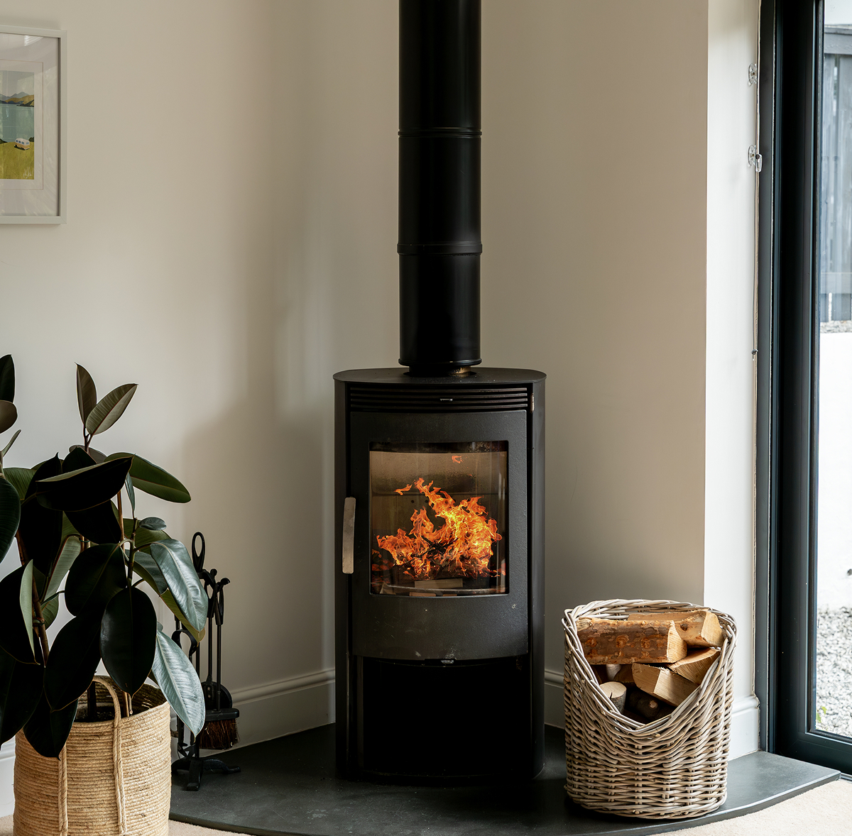 Cozy modern wood-burning stove with visible flames, a wicker basket of logs, and a potted plant in a bright room.