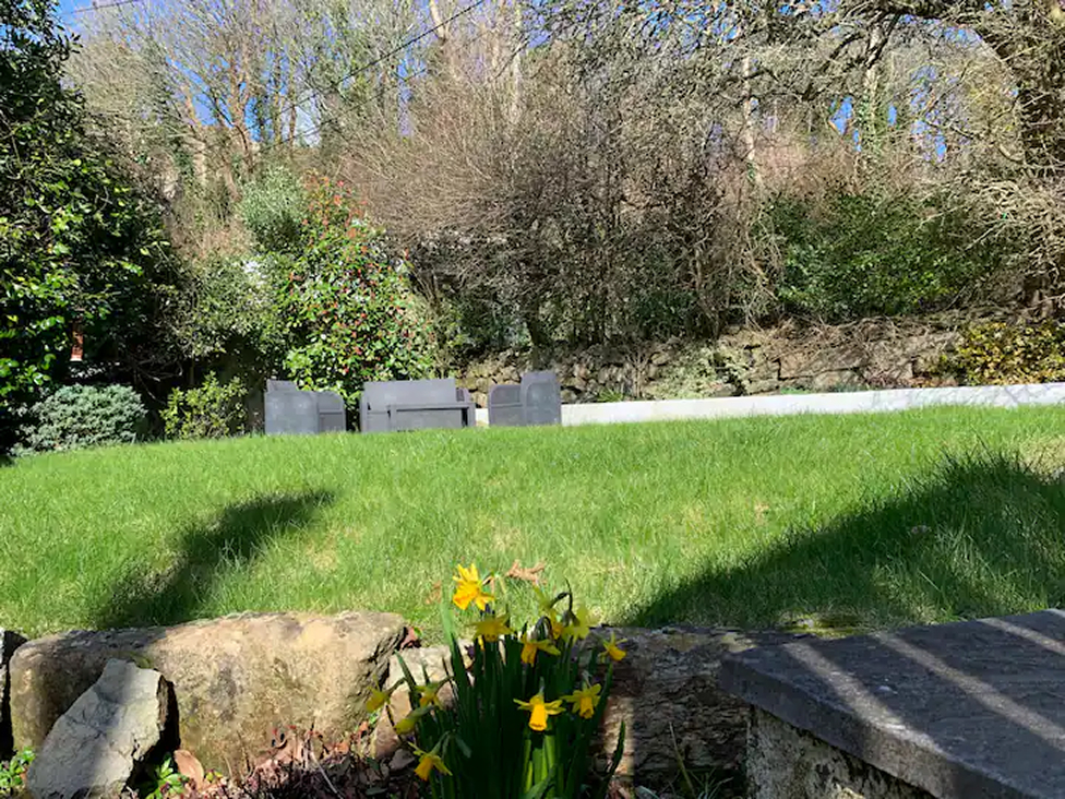 Sunny garden with yellow flowers in foreground, green lawn, gray patio furniture, and surrounding trees.